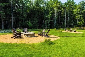 A campsite with a stone fire pit surrounded by Adirondack chairs, set in a grassy area with dense forest in the background.