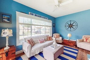 Un salon avec un mur bleu, un canapé blanc, une table basse et un panneau indiquant « PAS DE RÉSERVATIONS » placé au-dessus d’une fenêtre.