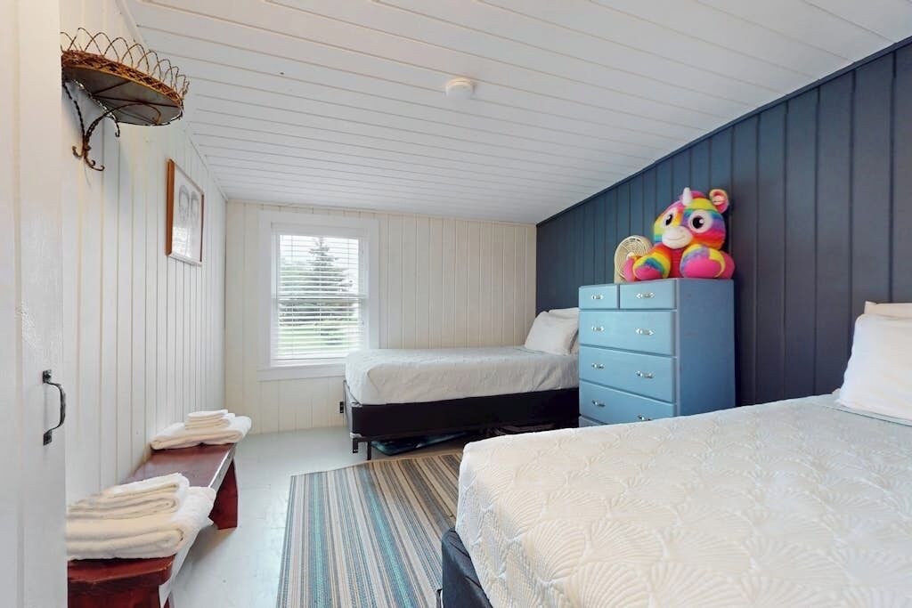 A bedroom with two beds, a blue dresser, a window with blinds, and a wall-mounted shelf.