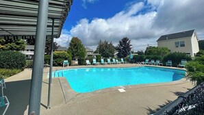En swimmingpool med liggestole, et overdækket område og en bygning i baggrunden.