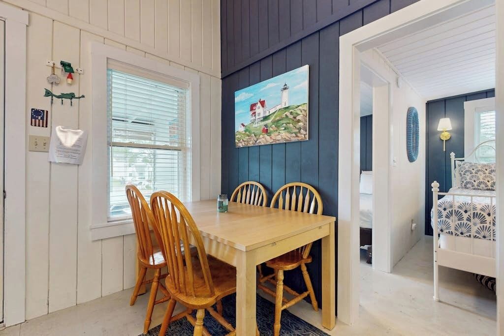 Un comedor con una mesa y sillas de madera, un cuadro de un faro, y una cama con una colcha estampada en la habitación contigua.