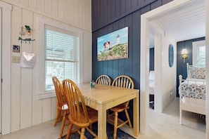 A dining area with a wooden table and chairs, a painting of a lighthouse, and a bed with a patterned bedspread in the adjacent room. - Cute cottage with screened porch & shared pool - walk to Ogunquit Beach (Wells)