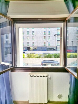 Ein Zimmer mit Fenster, Blick auf ein Gebäude mit vielen Fenstern und ein Auto vor der Tür.