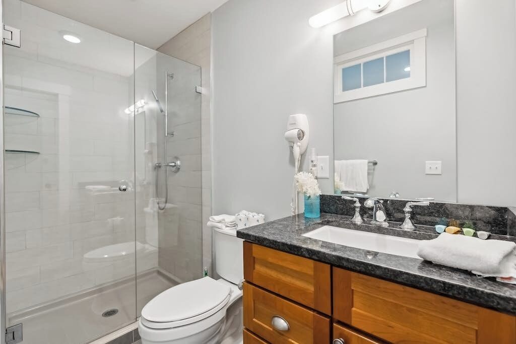 A modern bathroom with a glass-enclosed shower, a white toilet, a vanity with a sink, and a hairdryer on the wall.