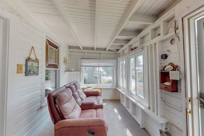 A room with a white wooden ceiling, a brown leather sofa, a window with a view, and a door with a shelf.