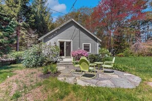 Ein Haus mit einer Steinterrasse, einer Haustür und einem kleinen Garten mit Bank und Stühlen.