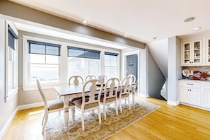 Un comedor con mesa y sillas, ventanas amplias y una cocina con armarios blancos.
