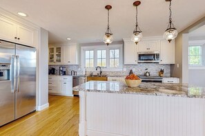 Una cocina con muebles blancos, un refrigerador de acero inoxidable, encimera de granito e iluminación colgante.