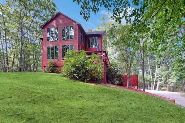 A two-story red house with a deck, surrounded by a well-maintained lawn and trees. - Charming Hillside Retreat with Hot Tub, Firepit, and Spacious Deck (Wells)