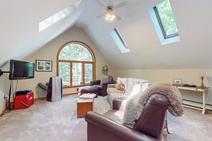 An attic room with a sloped ceiling, a large arched window, a ceiling fan, a television, a fireplace, and a desk with a lamp. - Charming Hillside Retreat with Hot Tub, Firepit, and Spacious Deck (Wells)