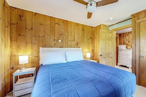 Quarto com paredes de madeira, cama com colcha azul, mesa de cabeceira com abajur e ventoinha no teto.