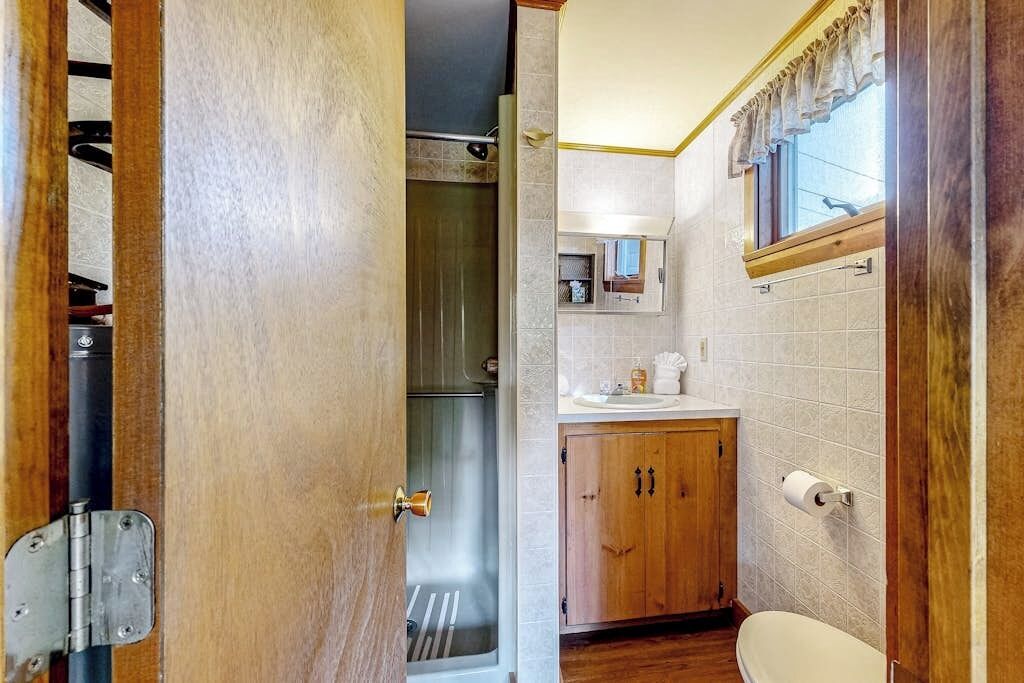 A bathroom with a wooden door, a shower, a toilet, and a small window with curtains.