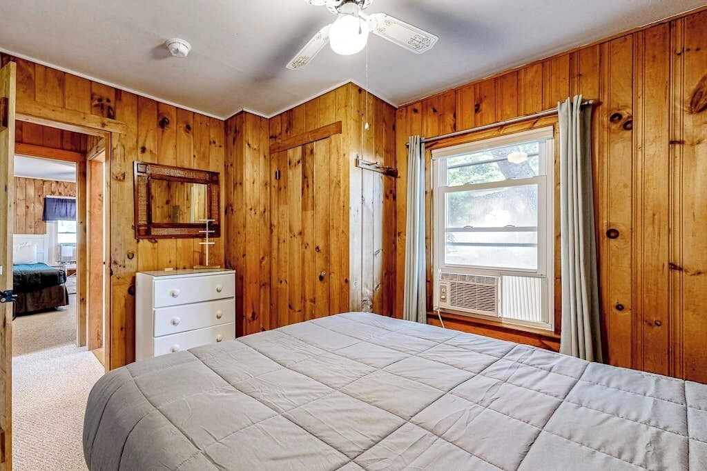 A wooden-paneled room with a bed, a dresser, a ceiling fan, and a window with curtains.