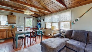 A cozy living room with a wooden ceiling, a sofa, a dining table with chairs, and a kitchen area with wooden cabinets and a stove. - Cozy Cottage with Private Deck - Walk to Short Sands Beach (York)