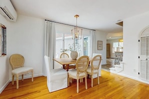 A dining area with a wooden table, four chairs, a chandelier, and a view through large windows.