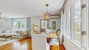 Uma sala de estar ampla com uma mesa de jantar de madeira, cadeiras estofadas brancas, um sofá e um candelabro.