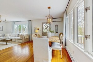 A spacious living room with a wooden dining table, white upholstered chairs, a sofa, and a chandelier.