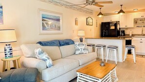 A living room with a sofa, a coffee table, and a kitchen area with a refrigerator and bar stools. - Coastal luxury condo steps from Wells Beach w/ AC, WiFi & full kitchen (Wells)