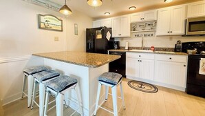 A kitchen with white cabinets, a black refrigerator, a granite countertop, and three bar stools. - Coastal luxury condo steps from Wells Beach w/ AC, WiFi & full kitchen (Wells)