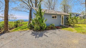 A small, single-story house with a gravel driveway, surrounded by trees and a grassy yard. - Inviting oceanfront home with fireplace, firepit, & views from every room (York)