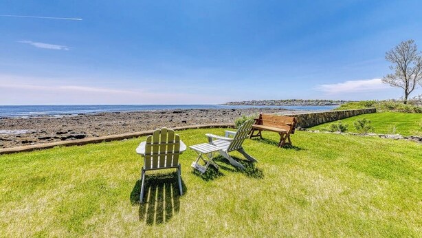 A grassy outdoor area with Adirondack chairs and a wooden bench, overlooking a rocky shoreline. - Inviting oceanfront home with fireplace, firepit, & views from every room (York)