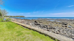A rocky shoreline with a grassy area and a clear blue sky. - Inviting oceanfront home with fireplace, firepit, & views from every room (York)