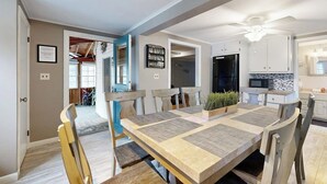 Uma sala de jantar com uma mesa comprida, cadeiras, um ventilador de teto e uma cozinha ao fundo.