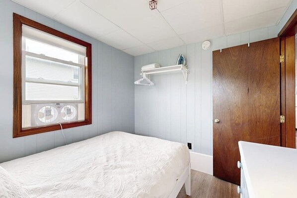 A bedroom with a single bed, a wooden door, a window with blinds, and a small shelf with a hanging garment.