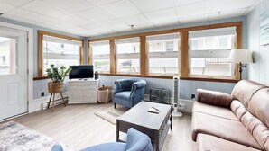 Una habitación con televisión, un sillón azul, un sofá marrón y ventanas amplias.