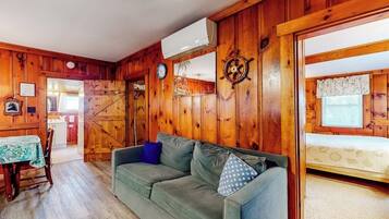 A room with wooden walls, a sofa, a dining table with chairs, and a bed with a window.