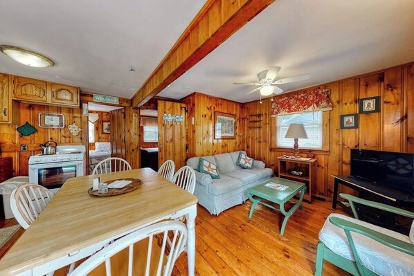 A wooden cabin interior with a dining table, chairs, a sofa, a television, and a ceiling fan.