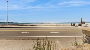 A coastal road with a beach in the background, a flagpole, and a small structure on the right.