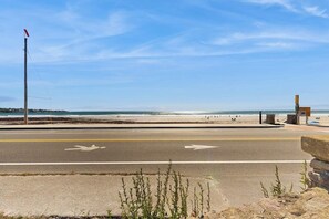 A coastal road with a beach in the background, a flagpole, and a small structure on the right.