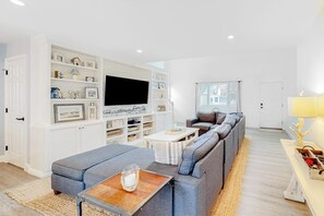 Ein modernes Wohnzimmer mit einem großen Flachbildfernseher, einem grauen Ecksofa, einem kleinen Couchtisch und einer Bücherwand mit Dekoration.