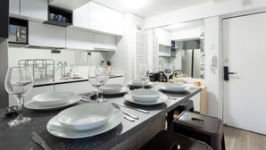 Uma cozinha moderna com armários brancos, bancada de mármore e espaço de jantar acomodando seis pessoas, com pratos, copos e talheres dispostos.