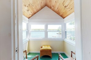 A room with a sloped ceiling, a bench with cushions, and a view of a body of water through large windows. - Charming lakefront cottage with a dock, WiFi, & washer/dryer - dogs welcome (Windham)