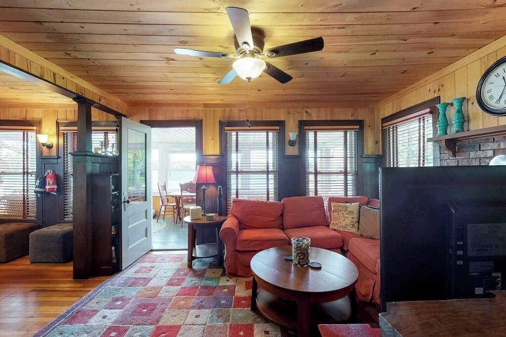 A cozy cabin interior with a wooden ceiling, a ceiling fan, a fireplace, a sofa, and a dining table.
