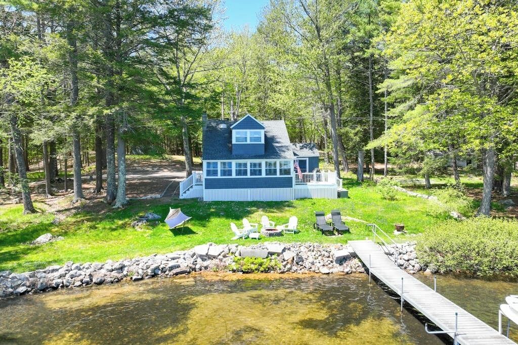 A cabin with a deck, surrounded by trees and a dock extending into a body of water.