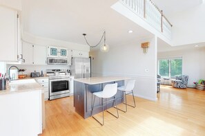 A modern kitchen with a central island, stainless steel appliances, and a dining area with a high ceiling and staircase. - Inviting home with ocean view, firepit, deck, golf nearby, & W/D (York)
