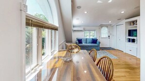 Un comedor con una mesa de madera, sillas y un cuenco decorativo. Se observa un sofá y un televisor al fondo.
