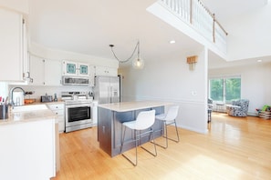 A modern kitchen with a central island, stainless steel appliances, and a dining area with a high ceiling and staircase. - Charming home with ocean view, firepit, deck, golf nearby, & W/D (York)