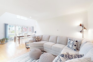 A modern living room with a sectional sofa, ottomans, and a dining area with chairs and a table. - Charming home with ocean view, firepit, deck, golf nearby, & W/D (York)