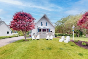 A two-story house with a porch, white chairs, and a tree with red leaves. - Charming home with ocean view, firepit, deck, golf nearby, & W/D (York)