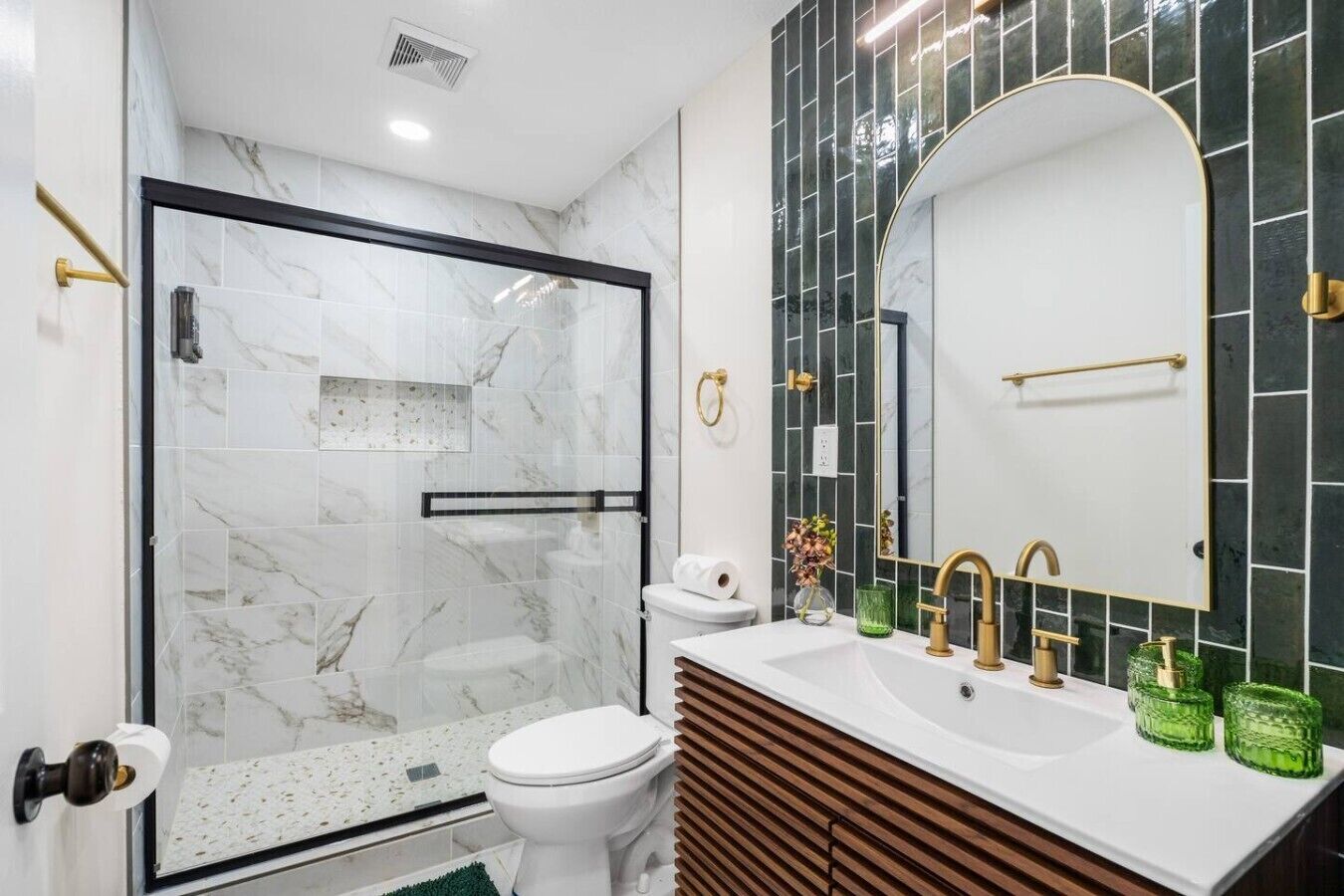 Une salle de bain moderne avec une douche à l’italienne en verre, des toilettes blanches, un meuble-lavabo avec un miroir et des porte-bougies en verre vert.