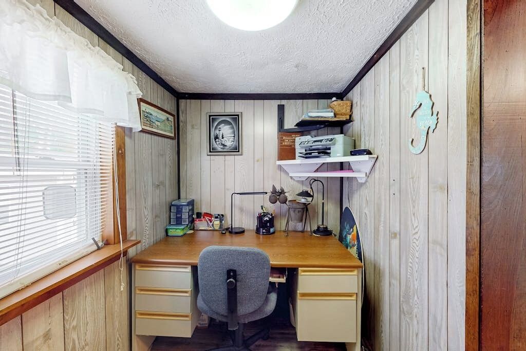 A room with a wooden desk, a chair, a window with blinds, a wall-mounted shelf with books, and a framed picture.