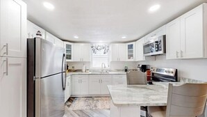 A modern kitchen with white cabinetry, a stainless steel refrigerator, a microwave, and a dining area with a marble countertop and chairs. - Adorable nautical home close to town, W/D, & streaming, dog friendly (Wells)
