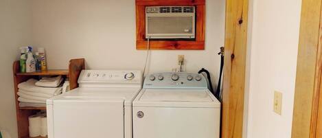 A laundry area with two stacked washers and dryers, a wooden shelf with folded towels, and a wall-mounted heater.