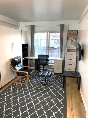 Ein kleiner, moderner Raum mit einem schwarz-weißen Musterboden, einem schwarzen Bürostuhl, einem Holzstuhl, einem Schreibtisch mit Stuhl, einem an der Wand montierten Fernseher und einem Fenster mit Vorhängen.