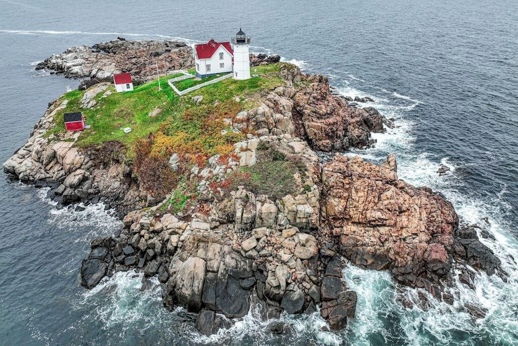 Ein Leuchtturm und ein weißes Gebäude mit rotem Dach auf einer felsigen Insel, umgeben von Wasser.