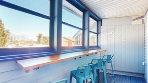 Un espace extérieur abrité, doté d’un banc en bois et de chaises en métal, de grandes fenêtres et offrant une vue sur les environs.